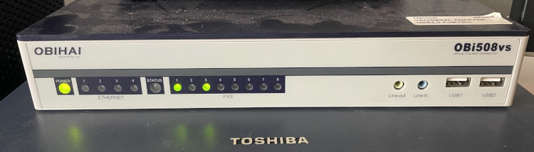 The device in question, a silver rectangle with a dark blue top, power, ethernet, status, fxs (the phone lines) status leds, two audio jacks and two USB ports.